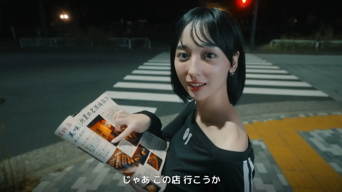 夜の街角で、一人の女性が雑誌を片手に笑顔で何かを指差しています。