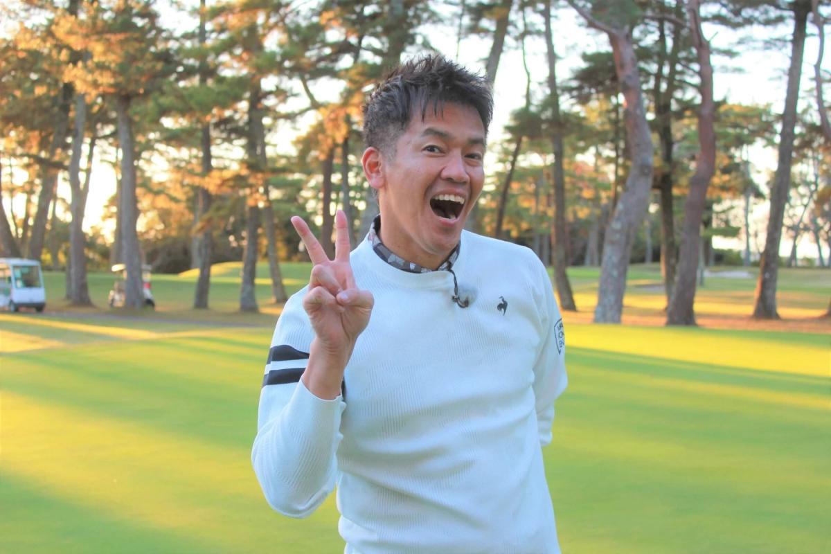 ゴルフ場で白いセーターを着た男性が、満面の笑みでピースサインをしている瞬間です。