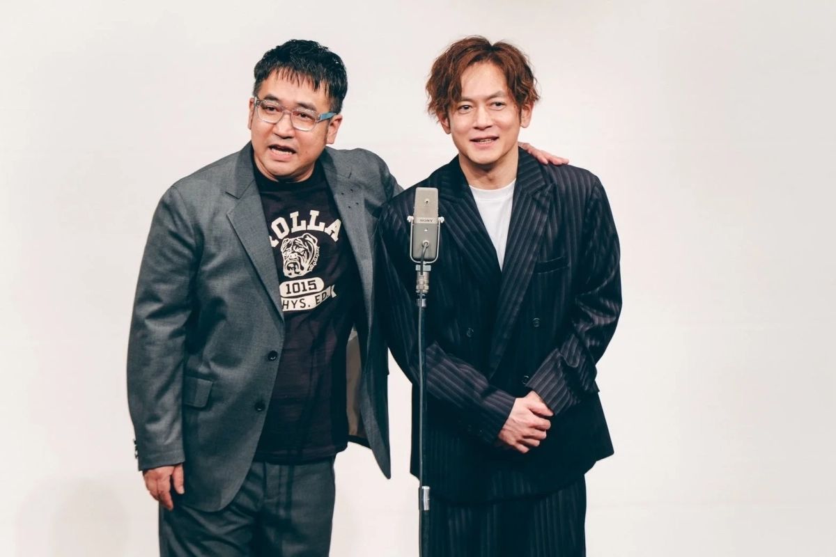 白い背景の前で、二人の男性がマイクの横に立っています。一人はグレーのスーツにTシャツ、もう一人はストライプのスーツを着ており、笑顔でカメラを見ています。