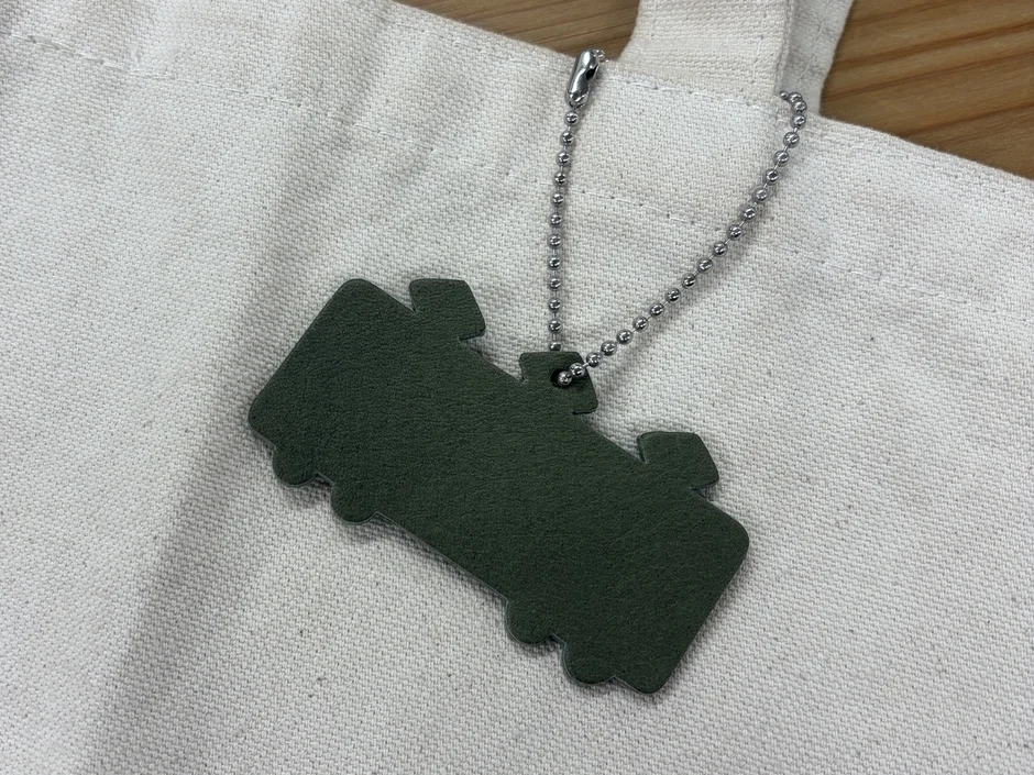 白いキャンバス地のトートバッグに、電車のような形をしたカーキ色のキーホルダーがボールチェーンで付けられている様子を上から撮影した写真です。シンプルで実用的な小物の組み合わせが特徴です。