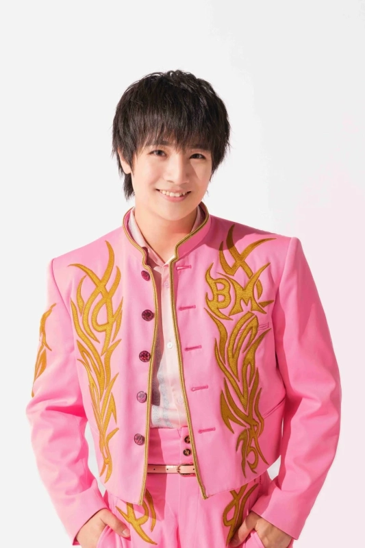 笑顔の若い男性が、金色の炎のような刺繍が施された鮮やかなピンクのスーツを着てポーズをとっている