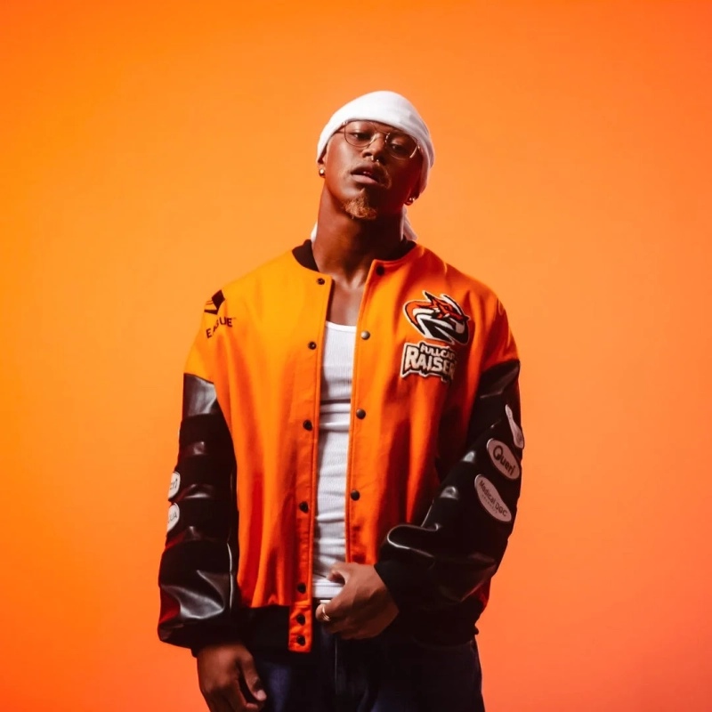 オレンジ色の背景をバックに、オレンジと黒のスタジャンと白いヘッドバンドを身につけた男性がポーズをとっています。眼鏡をかけ、スタイリッシュで自信に満ちた表情が印象的です。