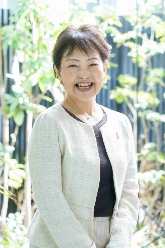 屋外の緑を背景に、白いジャケットを着た笑顔の中年女性のポートレート