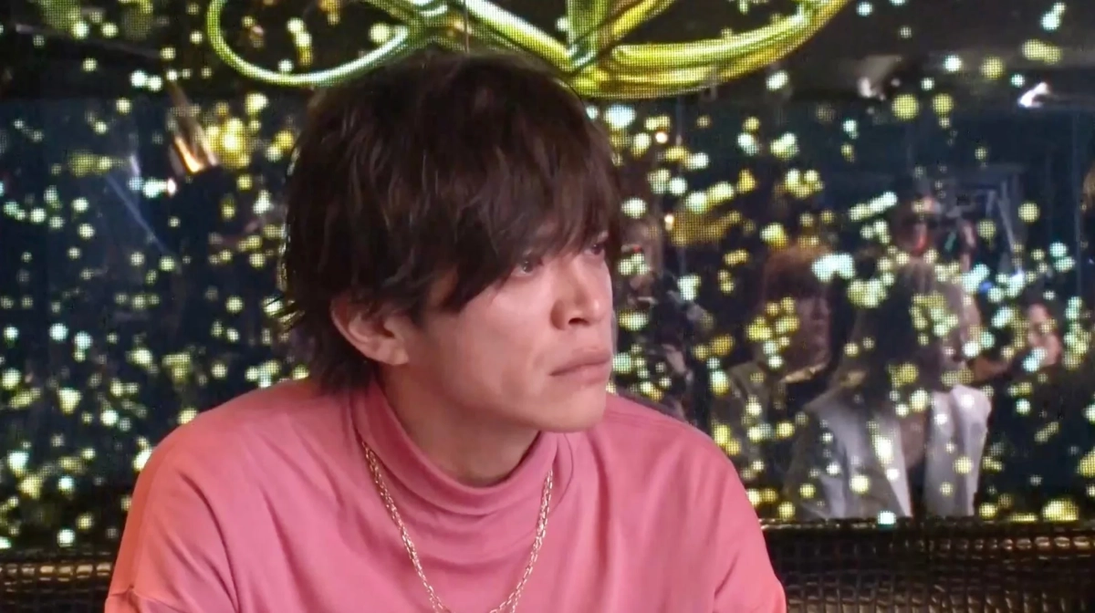 ピンクのタートルネックとゴールドのネックレスを身につけた男性が、真剣な表情で横を見ている様子