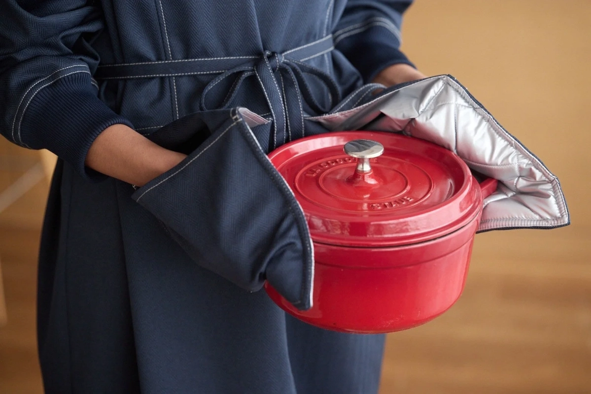 ネイビーのエプロンを着用した人物が、赤いストウブ鍋をオーブンミトンで持ち運んでいる様子を捉えた画像です。