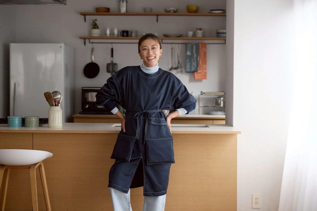 モダンなキッチンで笑顔で立つ女性のポートレート。紺色のエプロン風の服を着ており、清潔感のある明るい空間が広がっています。棚には調理器具や小物が整然と並べられています。