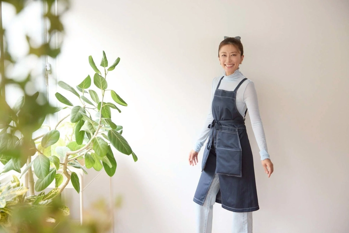 明るい室内で、ネイビーのエプロンを着た笑顔の女性が立っています。左手には大きな観葉植物があり、全体的に清潔感のある雰囲気です。