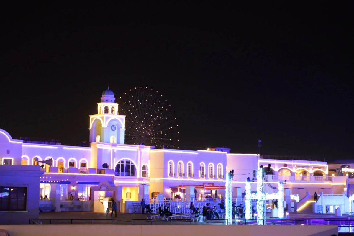 夜空の下、紫と黄色のネオンで美しくライトアップされたヨーロッパ風の建物群と時計台が印象的です。奥には観覧車が輝き、広場では人々が夜の雰囲気を楽しんでいます。