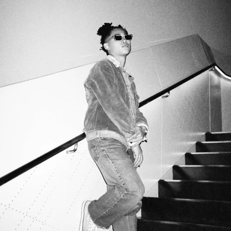 モノクロ写真で、サングラスをかけた人物が階段に立つ姿を捉えています。デニムジャケットとジーンズを着用し、両手を組んでクールな雰囲気を醸し出しています。屋内でのスナップショットのようです。