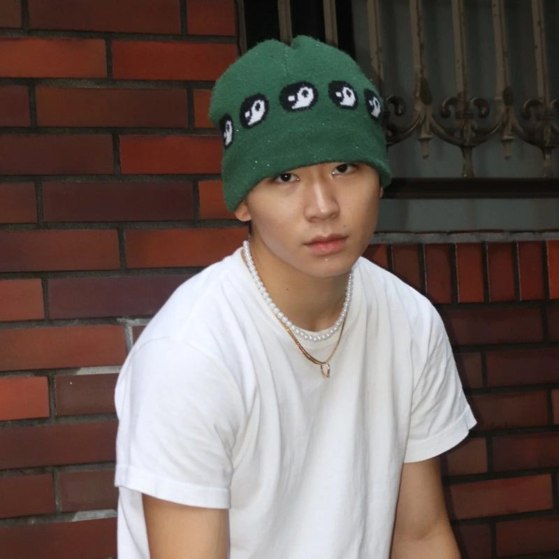 緑色のニット帽と白いTシャツを着用した若い男性が、カメラをまっすぐ見つめています。首元にはパールとゴールドの2連ネックレスを重ね付けしており、背景はレンガの壁です。