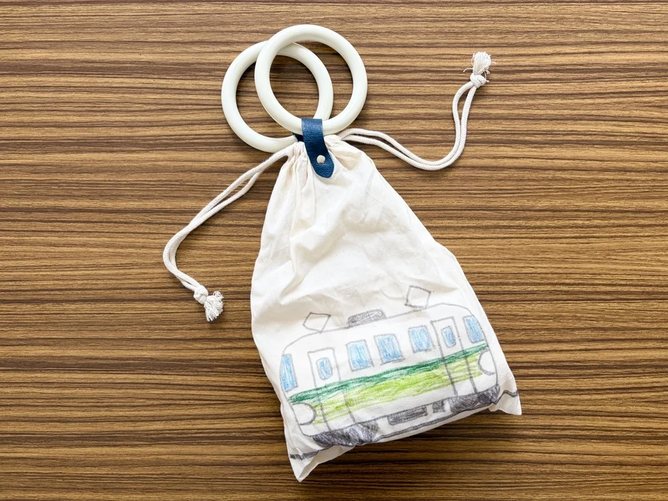 木製のテーブルの上に、子供が描いた電車の絵がプリントされた白い巾着袋が置かれています。白いリング状の持ち手が付いており、手作りの温かみを感じさせます。