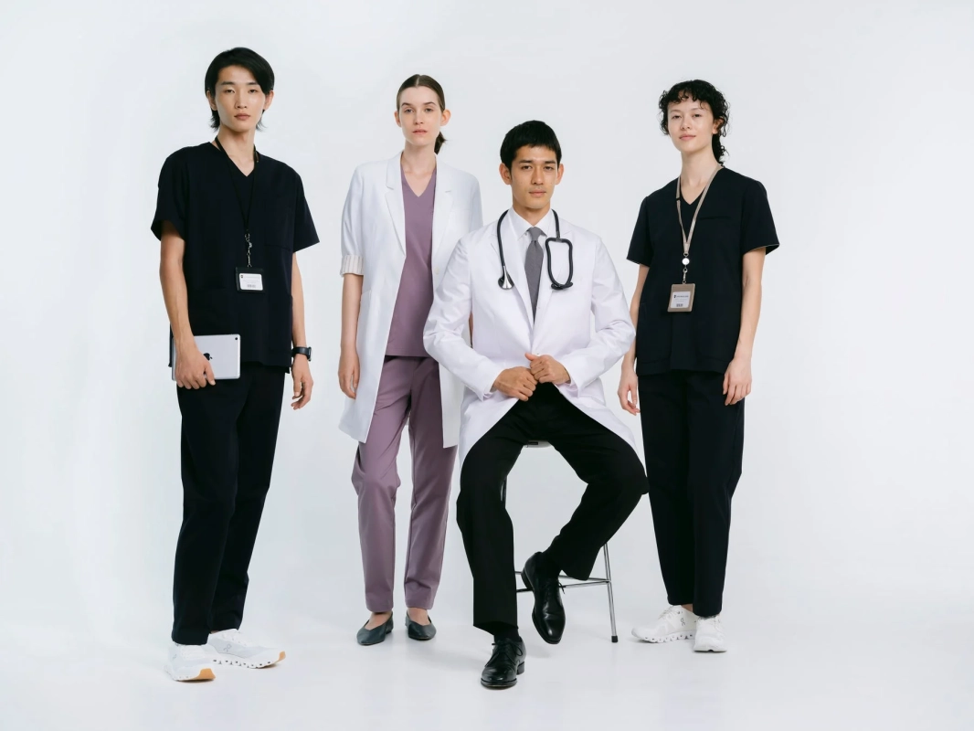 白い背景の前で、4人の医療従事者が様々な医療ユニフォームを着用してポーズをとっている。中央に座る男性医師と、その両脇に立つ男女のチームが、プロフェッショナルな雰囲気を醸し出している。