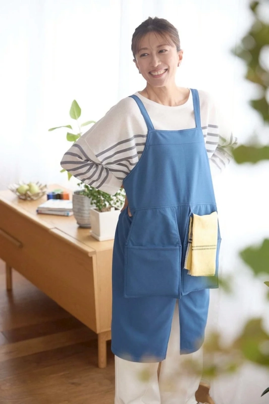 明るいリビングで、青いエプロンとストライプのシャツを着た女性が笑顔で立っています。両手を腰に当て、家事をする準備ができたような楽しそうな表情です。