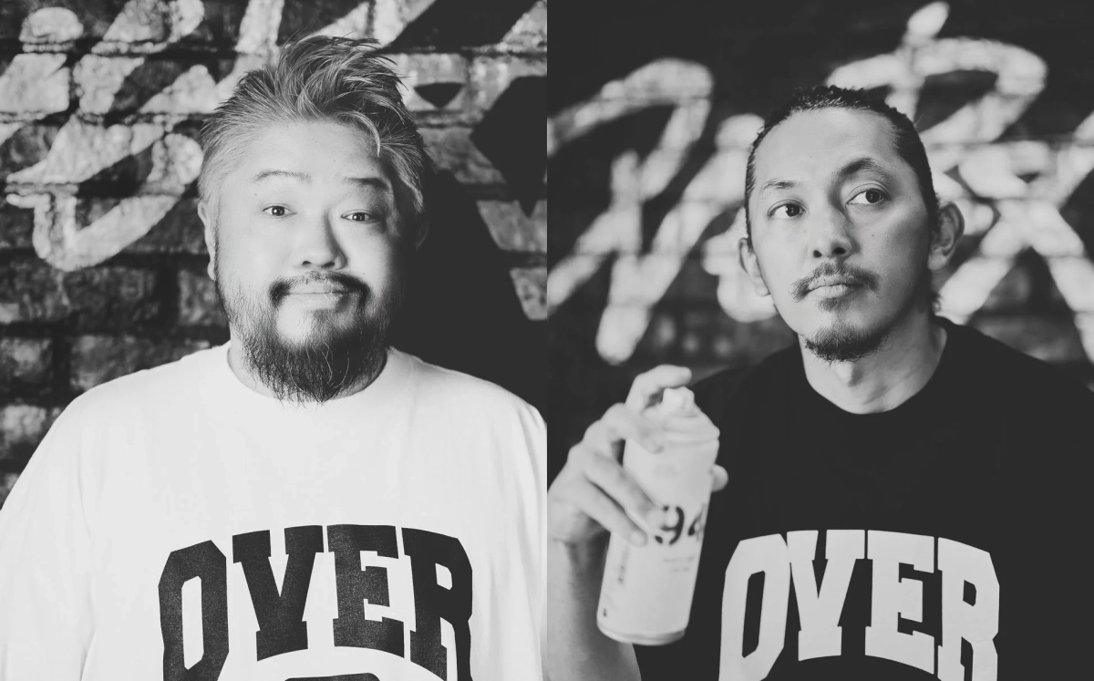 グラフィティが描かれたレンガの壁を背景に、2人の男性が並んで写る白黒写真。OVER ALLsの代表とアーティスト