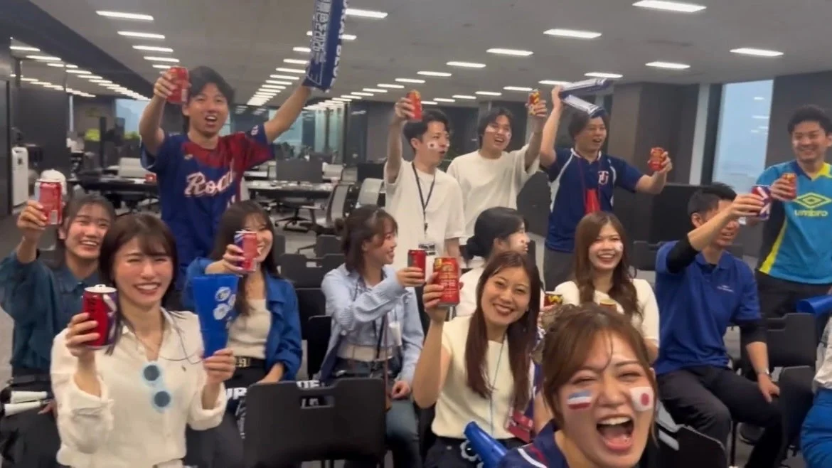 オフィスで大勢の若者たちが、飲み物を掲げて歓喜している様子