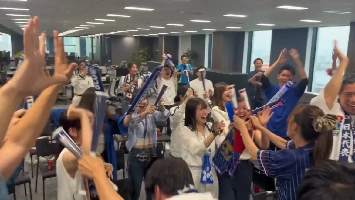 オフィスで多くの人々が日本代表のユニフォームやグッズを身につけ、熱狂的に応援している様子