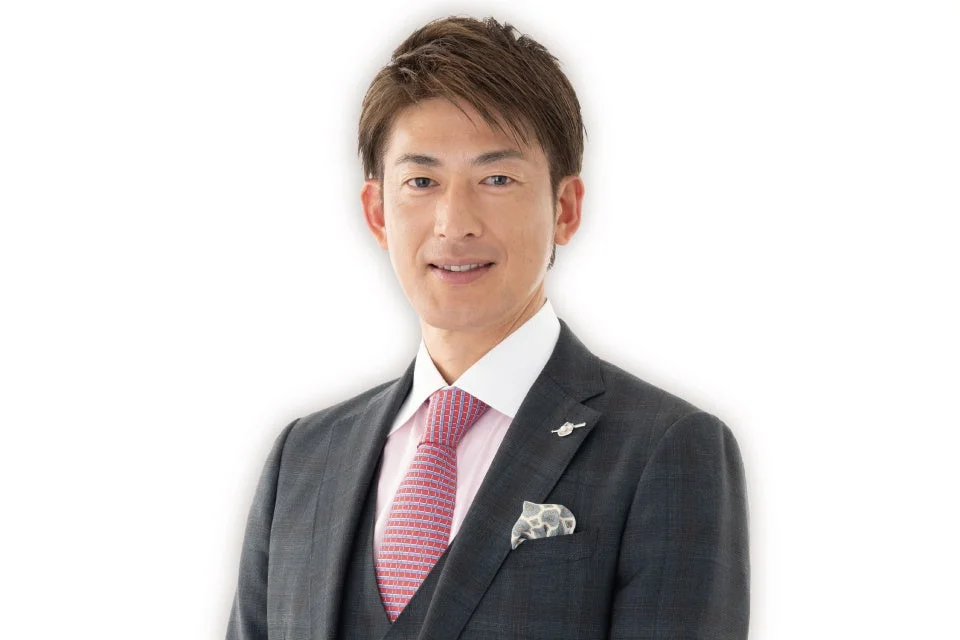 グレーのスーツとピンクのシャツ、赤のネクタイを着用した日本人男性が、穏やかな笑顔でカメラを見つめているポートレート