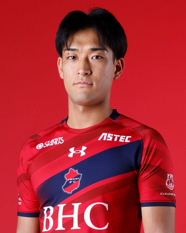 赤い背景の前で、赤いユニフォームを着用した若い日本人男性のポートレートです。ユニフォームには「IWAKI FC」のロゴや複数のスポンサー名が見られ、プロサッカー選手であると考えられます。
