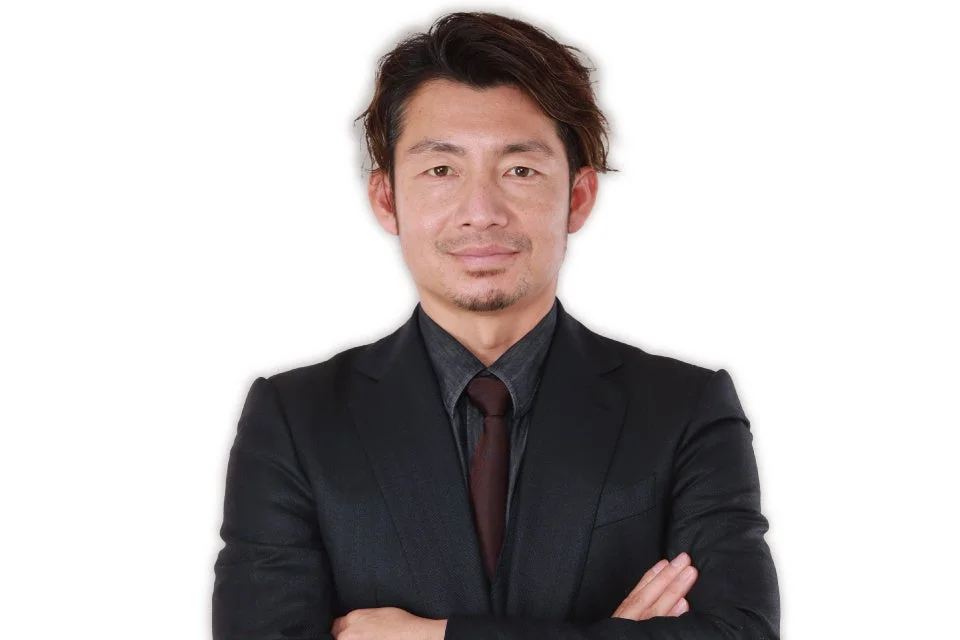 黒いスーツとシャツ、茶色のネクタイを着用したアジア系の男性が腕を組んで立っているポートレート写真