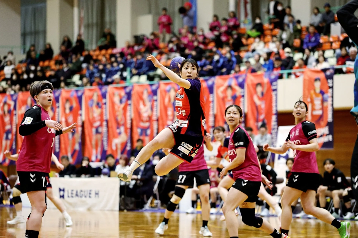 ハンドボールの試合中、赤いユニフォームの選手がジャンプしてボールを投げようとしている
