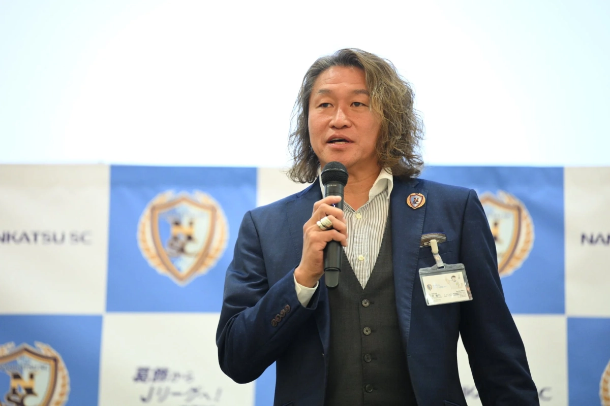 スーツ姿の男性（岡野雅行事業本部長）がマイクを手にスピーチ。背景に「NANKATSU SC」「夏からJリーグへ！」のロゴ。